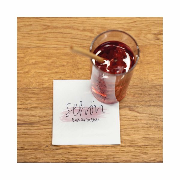 Weiße Mank Cocktailserviette „Schön, dass Du da bist!" neben einem Getränk